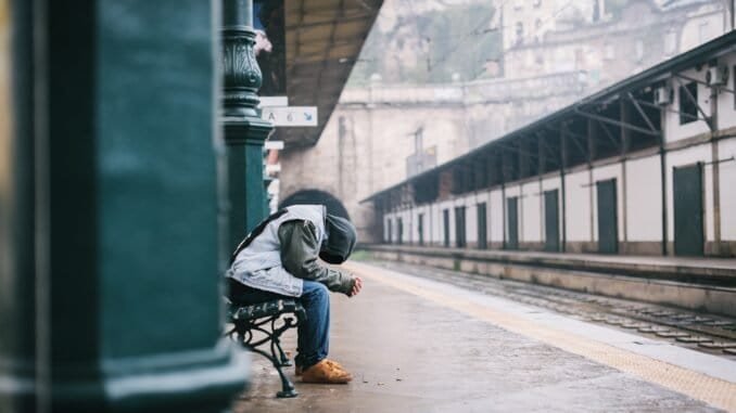 Homeless person sitting on a bench at a train station in cold weather.